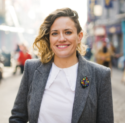 Person smiling outdoors in a city street, wearing a gray blazer.