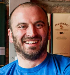 Smiling man with a beard wearing a blue shirt, standing indoors.