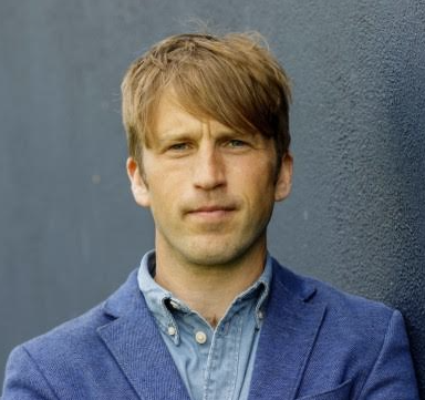Man in blue blazer standing against a dark wall.